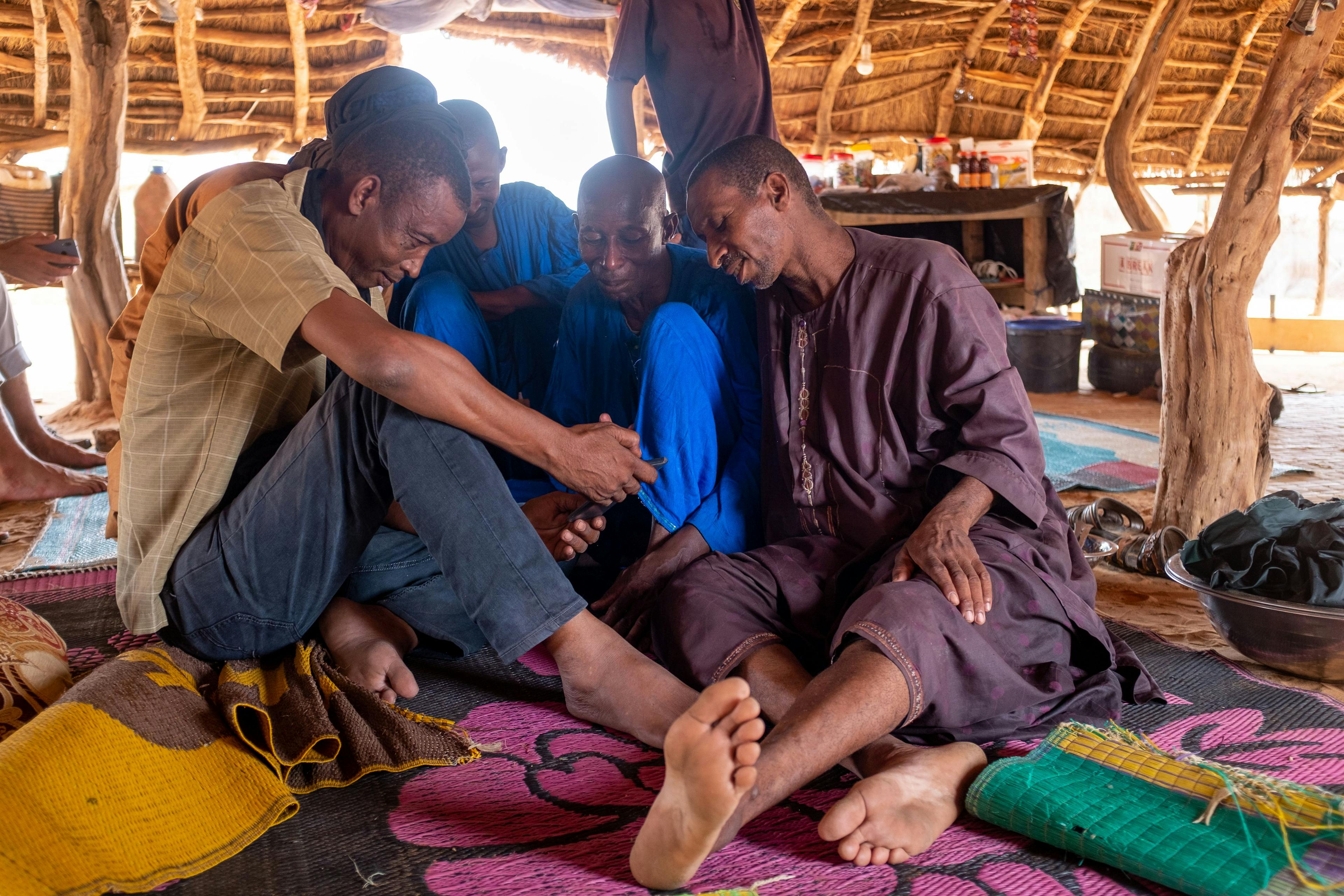 Fulani men huddle around a mobile phone to catch up on family news. As some herders use partial settlement — agro-pastoralism — to adapt to climate change, families become separated. 